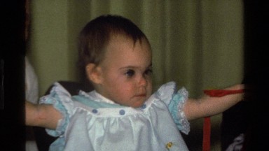 1977: baby in blue ruffled dress with ruffled pinafore, playing with ribbon