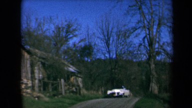1969: a car drives down a small hill past a large wooden house set in the woods