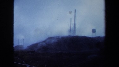 1971: clouds billow from large pipes sticking out of the ground on a high hill.