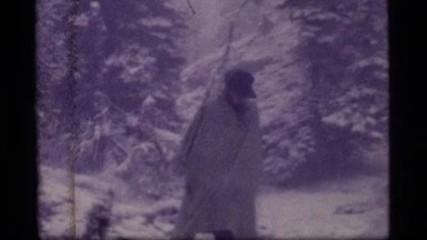 1977: Man Walking Through Snowy Forest Campsite Wrapped in Blanket