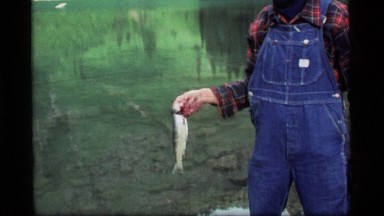 1977: Man Standing by Lake Holding a Trout by the Gills Sapphire Lake Montana
