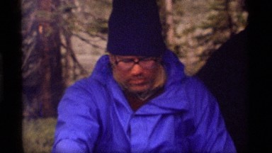 1977: a scruffy, rugged older man sits talking at a campfire SAPPHIRE LAKE