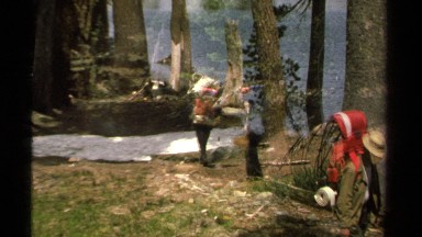 1978: hikers with large backpacks walk among trees by lake