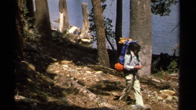 1978: hikers carrying backpacks hike on a forest trail near the bank of a lake