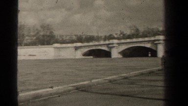 1947: a panning view of a river flowing under a bridge and near some buildings