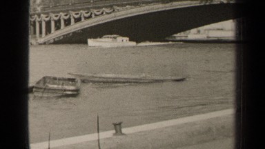 1947: Chugging Boat on Canal with Stone Bridge and City Skyline in Paris, France
