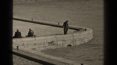 1947: man standing on a sea wall with a fishing pole; other men sitting on wall