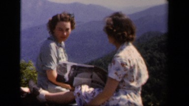 1943: Two Women Look at Brochure While Sitting on Ledge with Mountain Views