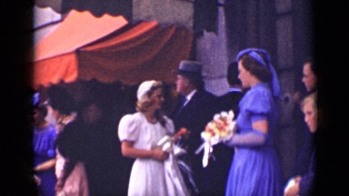 1940: women in fancy dresses standing in front of building holding flowers.
