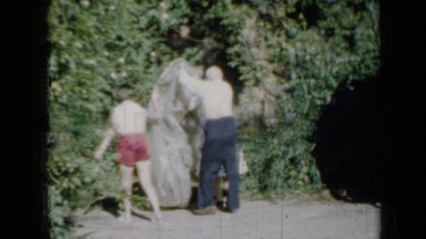 1958: older man and two girls move plastic cover and green car is by garage
