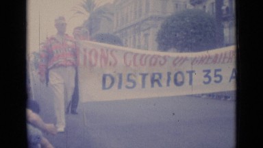 1962: people gathered on street for parade as people in parade continue down
