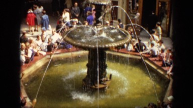 1971: tourists gather around european water fountain in commercial district.