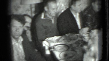 1975: Gathering of Men Finishing Meal Around Long Table in Small Crowded Room