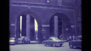 1962: cars travel through city archway while pedestrians wait to cross road