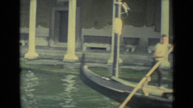 1962: Man Poles Boat Through Canal Next to Ancient Buildings
