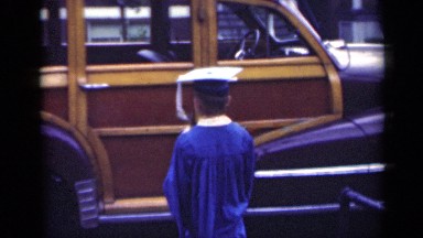 1948: boy in graduation cap walks amongst vintage cars and waves to the camera.