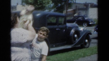 1950: large woman lifts and hugs infant in her arms with boy's help near car