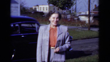 1950: well dressed lady smiles and talks to camera in front of vintage cars.
