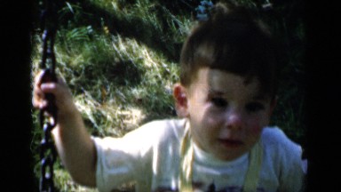 1951: small boy in yellow coveralls swings on a swing and smiles at the camera.