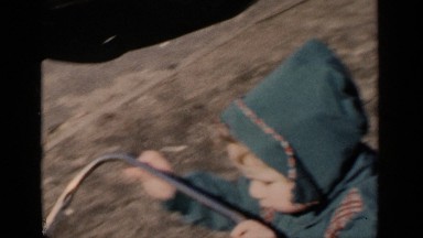 1951: toddler in a coat pushing a cart down a road outside, near a large city