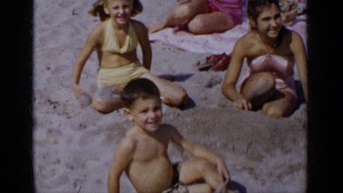 1951: woman smokes and folds garment while children play with beach sand