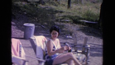 1964: woman waves while lounging and sunbathing on chair in sandy area GREENWOOD