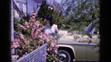 1965: well dressed woman in glasses stands in front of new car in her driveway.