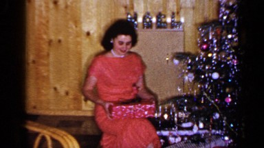 1965: woman wearing orange dress sits and unwraps present by christmas tree