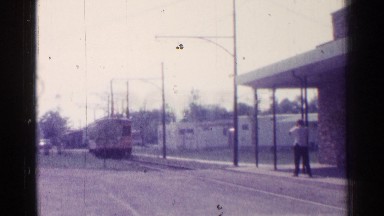 1976: man waits under awning at station for a yellow electric tram. ILLINOIS