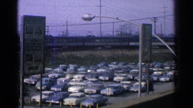 1965: train is moving in the rail where more and more cars are parking near