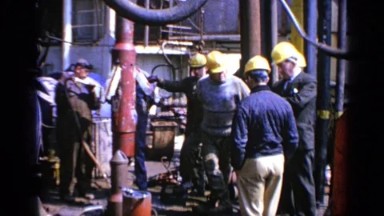 1970: Group of Men Wearing Yellow Hard Hats Standing Around a Long Pipe