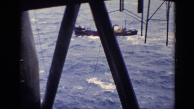 1970: a black barge moving through the sea under a huge metallic structure