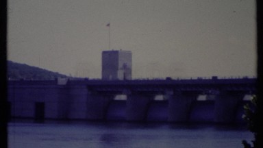 1971: bridge and building over water in front of a hill with cars driving