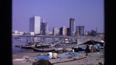 1970: people sitting on the sand looking at parked boats in the water. INDIA
