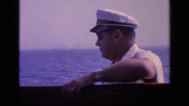 1968: a sailor on a boat walks gingerly while his son smiles at the camera.