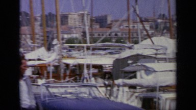 1963: Woman in White Dress on Ocean Bank with Parked Boats and Car, Greece