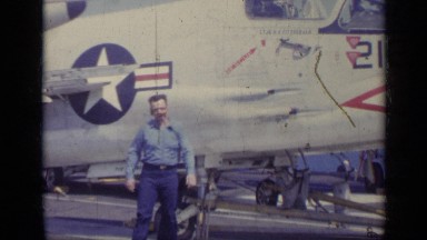 1962: man walks toward camera in front of a fighter plane aboard a navy ship.