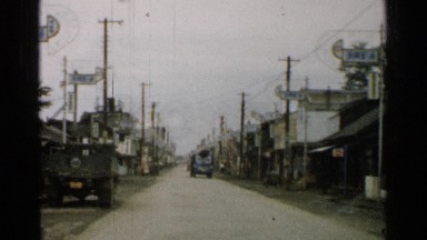1952: driving down a street with other cars and buildings and shacks lining