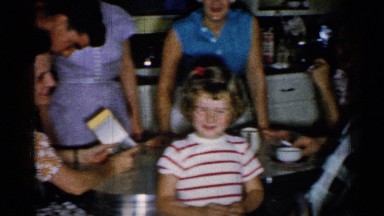1952: little girl smiling in a kitchen full of people and a man waving over her