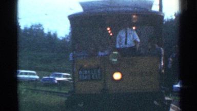 1953: people riding a tram, conductor hanging from the side, cars parked