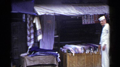 1953: sailor stands in front of outdoor stall displaying bolts of fabric JAPAN
