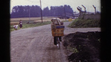 1953: a man riding in a bicycle in the road with heavy things loaded in that,