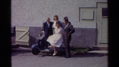 1961: a lady in a white dress sitting on a scooter posing with other people.