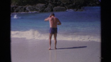 1967: man wearing blue swim trunks walks toward low waves and stops US VIRGIN