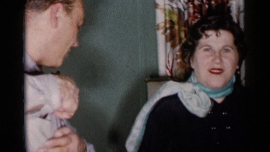 1955: man and woman standing in living room sit on a brown sofa and wave