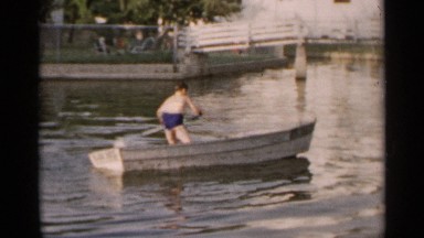 1954: young boy paddling a boat across the still water in blue swimming shorts