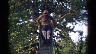 1954: two shirtless adult men slide down a child's slide at a park. NEW YORK