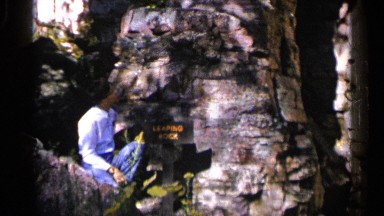 1960: man sitting down on a rock next to a sign with yellow writing on it. SIOUX