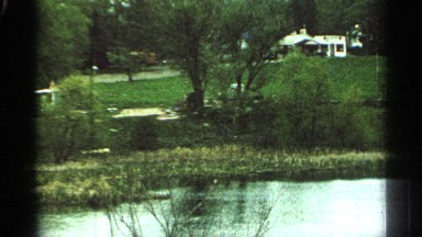 1965: a view of a park next to a creek at daytime, with houses in the background