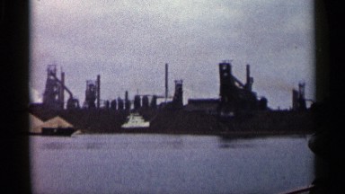 1960: man views industrial factories across a river while traveling on a boat